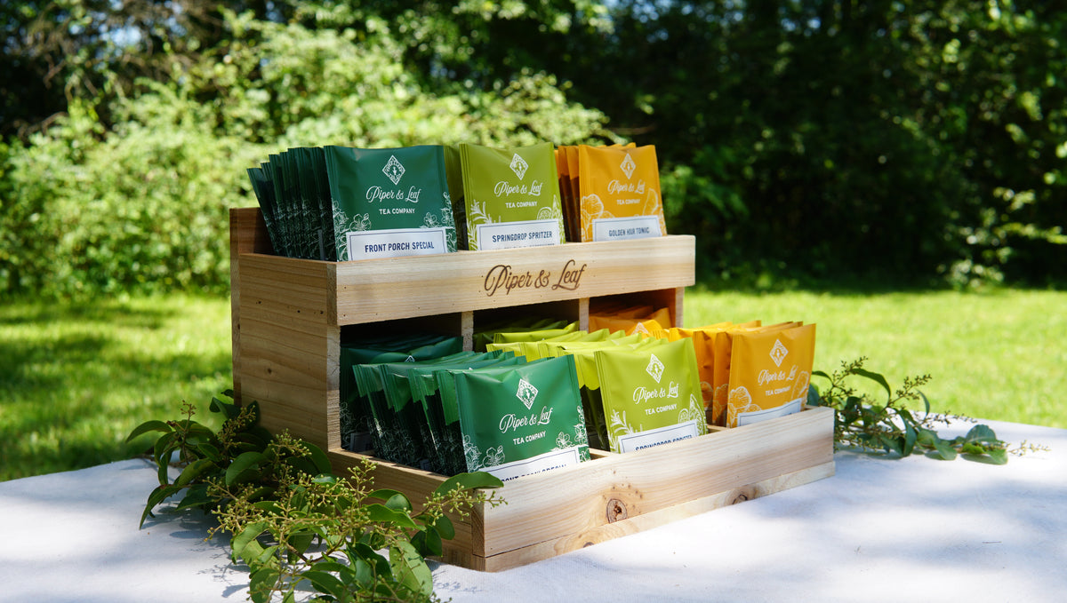 A Cedar Nook Display by Piper & Leaf Tea Co. holds assorted green, yellow, and orange tea bags outside on a white table with lush greenery—ideal for enjoying an al fresco tea tasting.