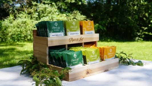 A Cedar Nook Display by Piper & Leaf Tea Co. holds assorted green, yellow, and orange tea bags outside on a white table with lush greenery—ideal for enjoying an al fresco tea tasting.