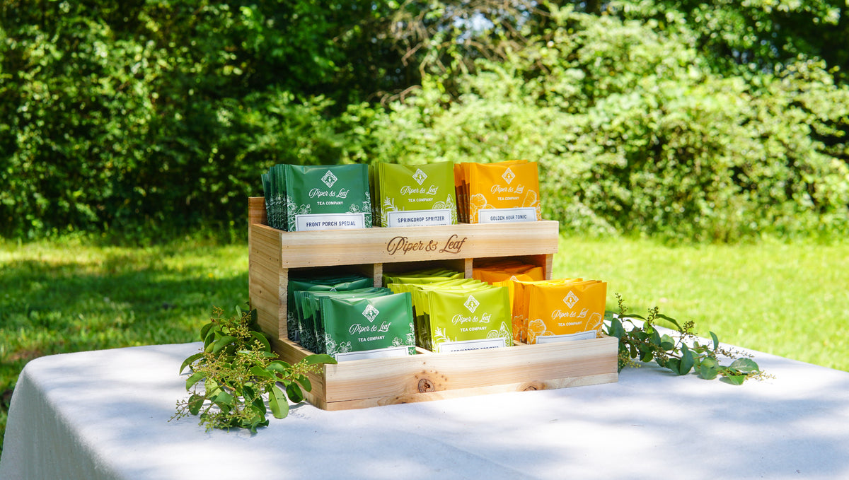 The Cedar Nook Display by Piper & Leaf Tea Co. shows off green and yellow tea bags on a white tablecloth outdoors, surrounded by lush greenery—an inviting setup for a tea tasting flight in nature.