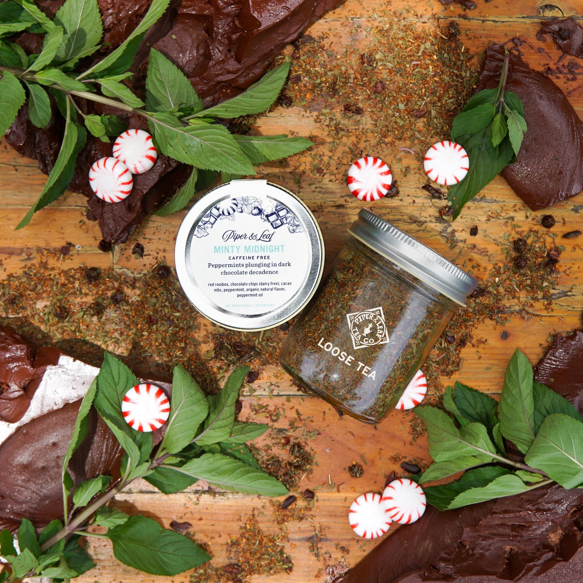 Two jars of Piper & Leaf Tea Co. Minty Midnight Glass Jar of Loose Leaf (30 servings) are displayed on a wooden surface with chocolate pieces, fresh mint sprigs, and peppermint candies scattered around.