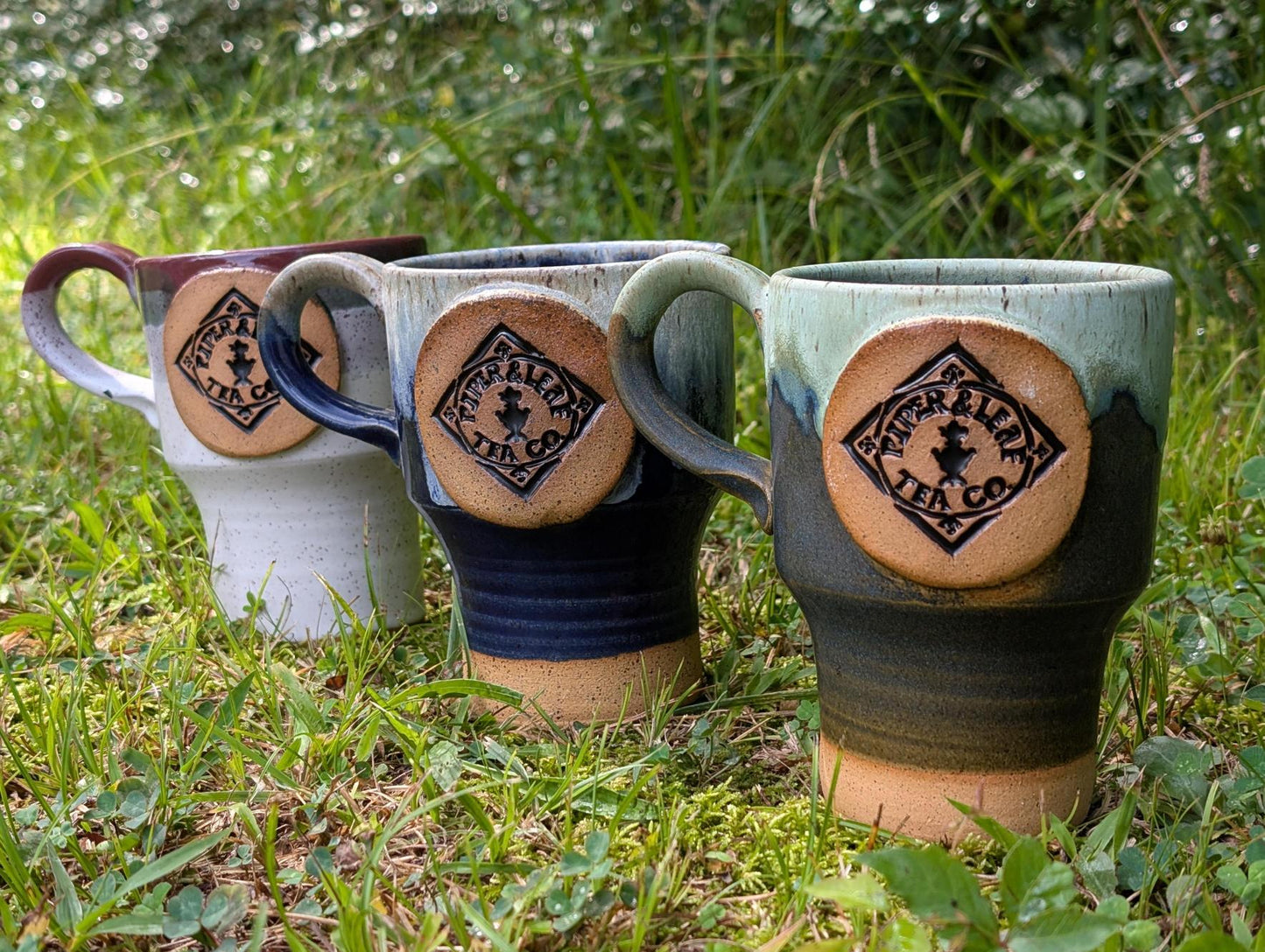 Three handmade Pottery Travel Mugs by Piper and Leaf Tea Co. sit on grass, arranged diagonally. The mugs are white, dark blue, and light green, with grass and greenery in the background.