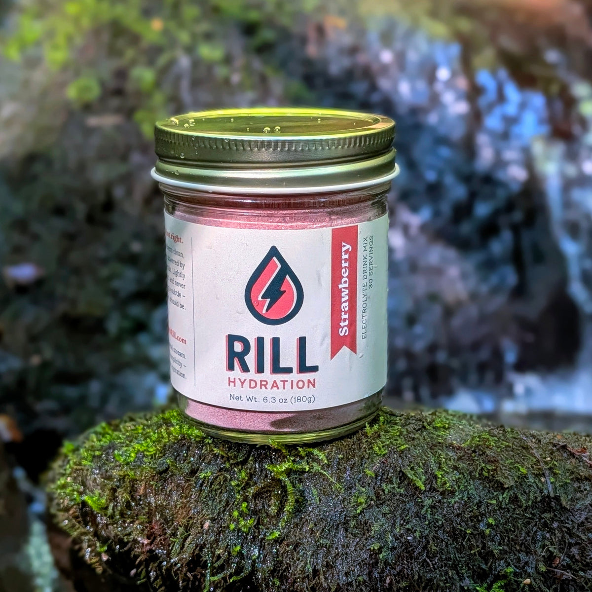 A jar of Rill Hydration’s Strawberry Rill Clean Hydration Drink Mix (30 serving bulk jar) with real fruit rests on a mossy rock, set against a blurred natural backdrop.
