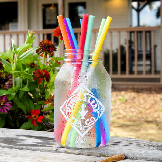 An UnStraw jar adorned with colorful, BPA-free straws from Piper & Leaf Tea Co. sits on a wooden table next to vibrant flowers.