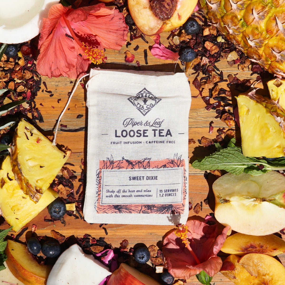 A Sweet Dixie muslin bag of loose leaf tea by Piper & Leaf Tea Co., with pineapple, peach, and hibiscus mingling on a wooden surface, creates a delightful fruit blend. Perfect for a refreshing, caffeine-free summertime brew.