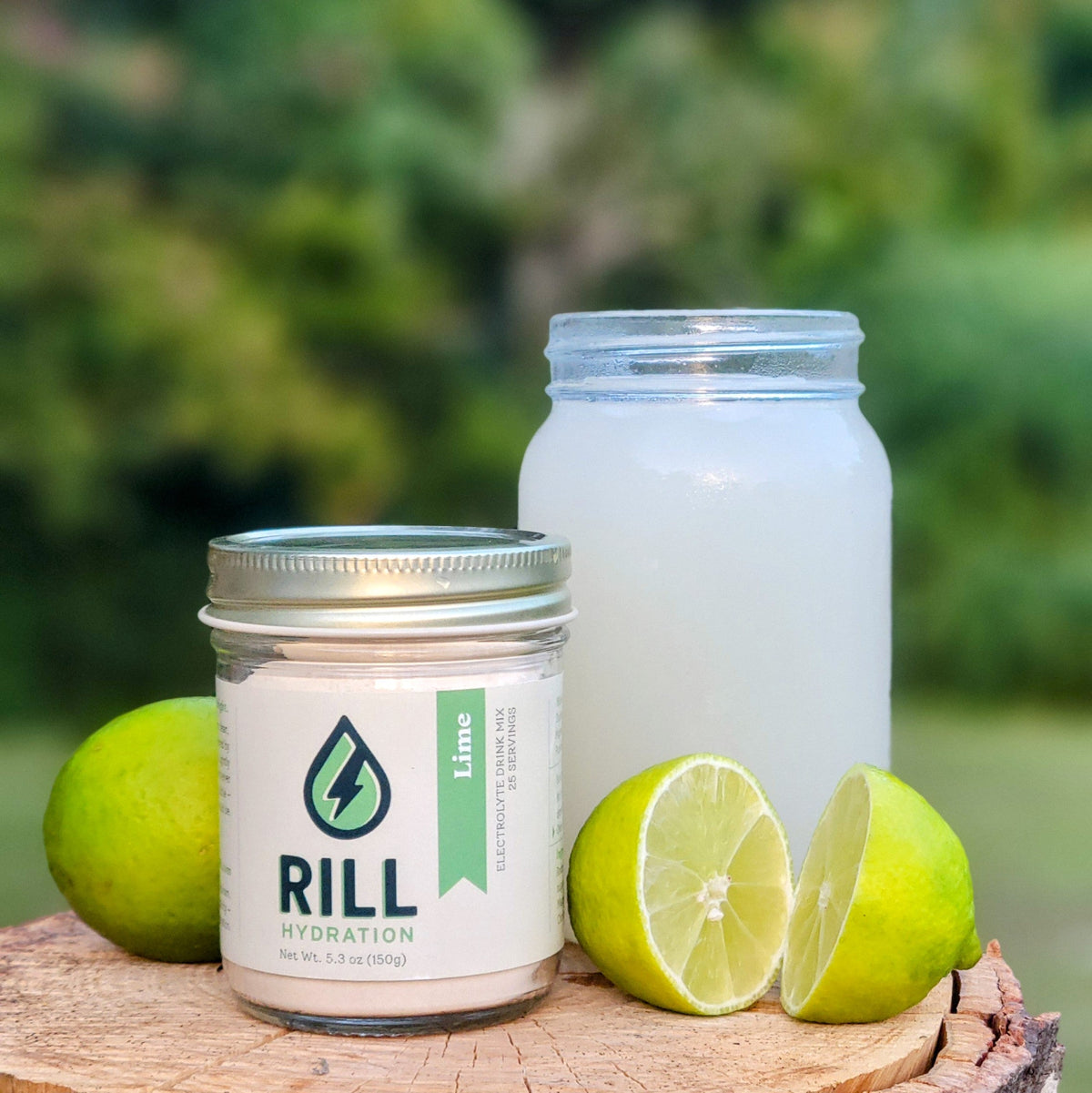 A jar of Rill Hydration’s Lime Rill Clean Hydration Drink Mix (30 servings), a glass of lime drink, and fresh limes—made with real fruit—are displayed on a wooden surface outdoors against a green backdrop.