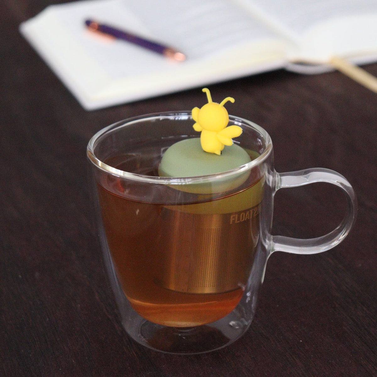 A clear glass mug of tea holds the Float2infuse Tea Infuser—Bumble Bee, a whimsical loose-leaf accessory topped with a small yellow bee and designed to float while steeping; an open notebook and pen in the background accentuate the charm of this delightful Floating Tea Infuser.